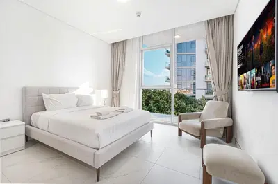 a white bedroom with a bed and a chair and a window at Elegant 1 Bedroom Apartment with Balcony & Lagoon in District One in Dubai