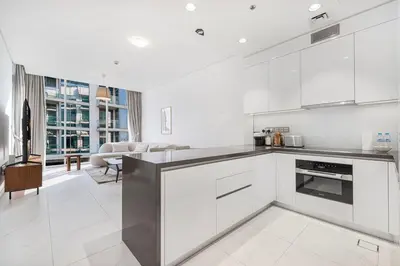 a kitchen with white cabinets and a living room at Elegant 1 Bedroom Apartment with Balcony & Lagoon in District One in Dubai