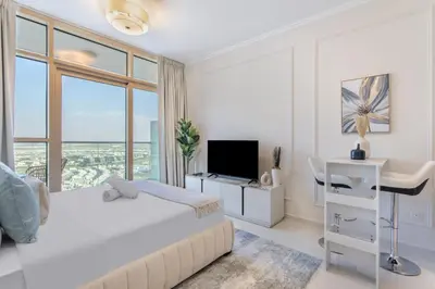 a white bedroom with a bed and a desk with a television at Sky-High Luxury Studio Overlooking Trump Golf Club-Carson A, DAMAC Hills, Dubai in Dubai